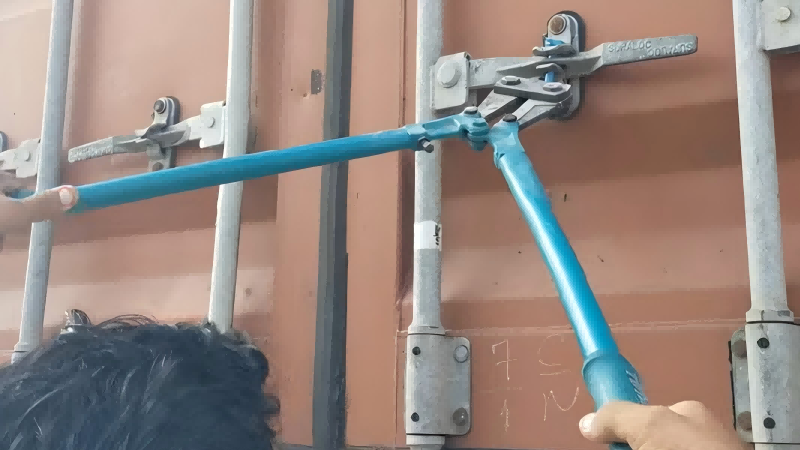 A close-up shot of a large pair of bolt cutters with its jaws positioned to cut through a high-security bolt seal on a container.