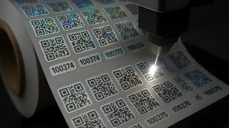 A laser marking system etching a unique QR code and serial number onto a roll of hologram stickers.