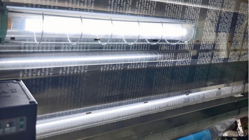 A massive industrial embossing machine with a heated roller pressing a pattern onto a continuous roll of silver film.