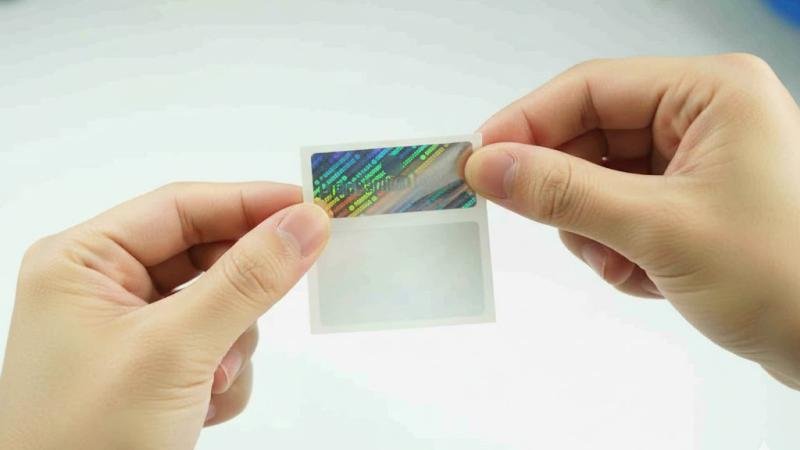 Fingers holding a hologram sticker by its edges, with the backing paper being peeled away.