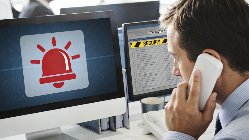 A manager on the phone, looking at a computer screen that displays a red alert icon next to a tracking entry for a security seal.